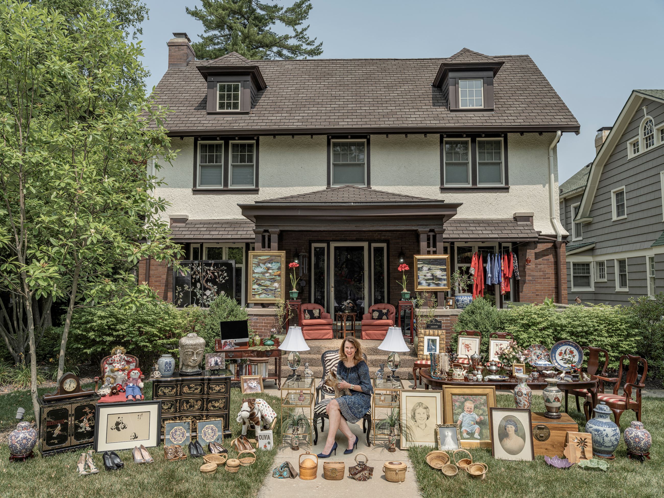 Photograph of a woman sitting in front of a house surrounded by household items.