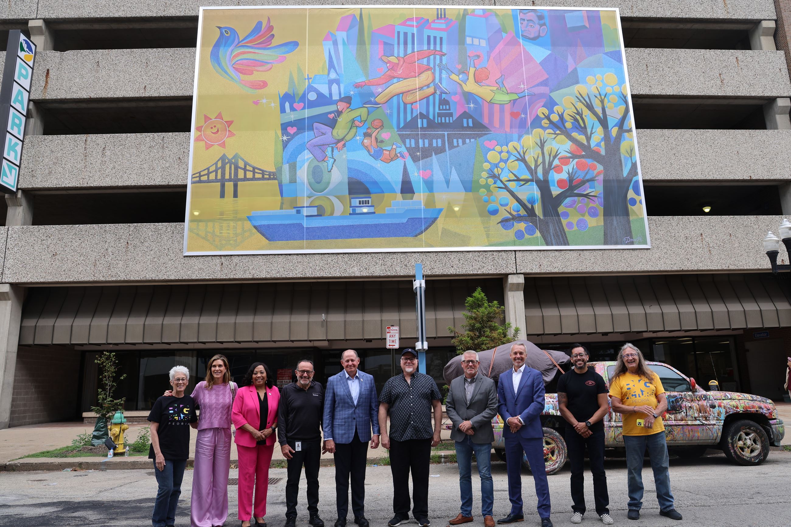 Row of people stand in front of a parking deck with a mural on it depicting play in Peoria.